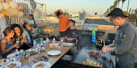 A chef in a grey uniform grilling meat and vegetables on a large flat-top hibachi grill for a group of people seated at a table on a rooftop.