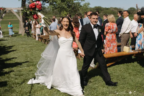 A bride holding a bouquet and a groom in a tuxedo walking down the aisle at an outdoor wedding ceremony.