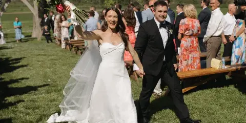 A bride holding a bouquet and a groom in a tuxedo walking down the aisle at an outdoor wedding ceremony.