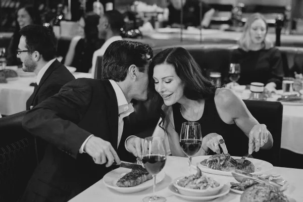 A couple enjoying a steak dinner in an upscale restaurant with wine glasses and other diners in the background.