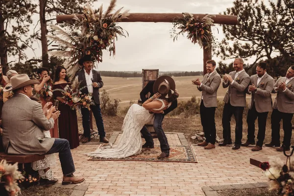 A bride and groom share a kiss during an outdoor wedding ceremony under a rustic wooden arch decorated with flowers, surrounded by guests.