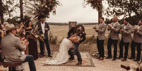 A bride and groom share a kiss during an outdoor wedding ceremony under a rustic wooden arch decorated with flowers, surrounded by guests.