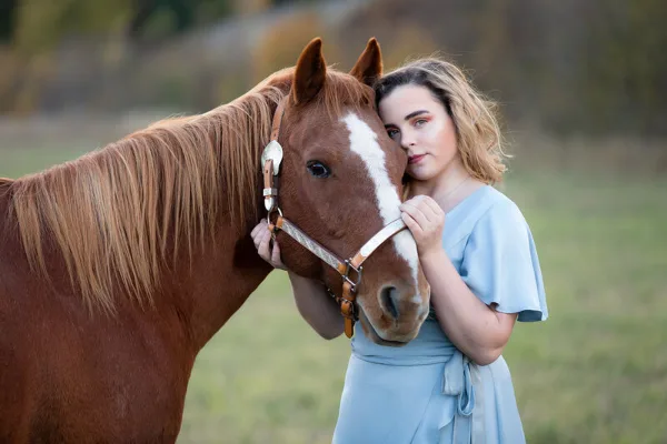 A young woman in a light blue dress posing affectionately with a brown horse in a field.