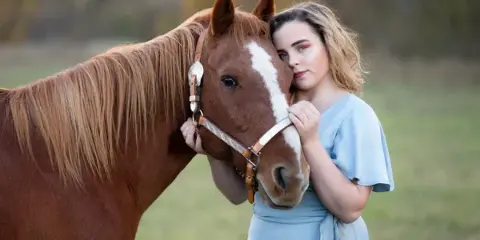 A young woman in a light blue dress posing affectionately with a brown horse in a field.