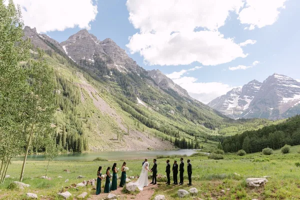 A bride and groom exchanging vows with their wedding party in a grassy field with a large mountain range in the background.