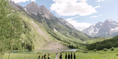 A bride and groom exchanging vows with their wedding party in a grassy field with a large mountain range in the background.