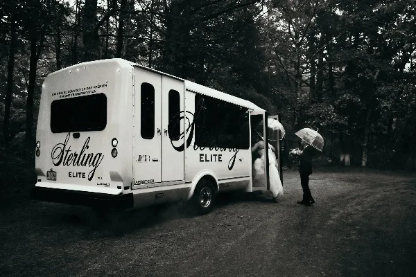 A white Sterling Elite shuttle van parked outdoors with a bride and groom standing near the open door holding umbrellas.
