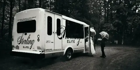 A white Sterling Elite shuttle van parked outdoors with a bride and groom standing near the open door holding umbrellas.