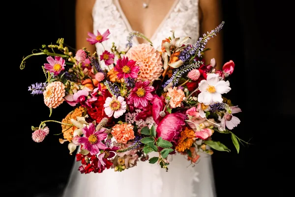 A bride in a white lace dress holding a large, colorful bouquet of mixed flowers including dahlias, cosmos, and foliage against a black background.