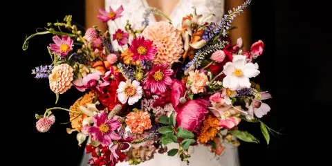 A bride in a white lace dress holding a large, colorful bouquet of mixed flowers including dahlias, cosmos, and foliage against a black background.