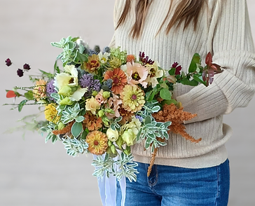 A person holding a large, colorful bouquet of seasonal cut flowers including dahlias, zinnias, and greenery.