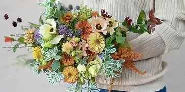 A person holding a large, colorful bouquet of seasonal cut flowers including dahlias, zinnias, and greenery.