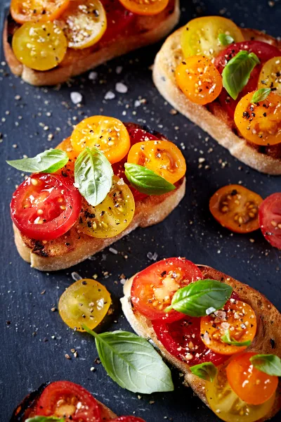 A close-up overhead shot of several pieces of bruschetta topped with sliced heirloom tomatoes, fresh basil leaves, and a sprinkle of coarse salt and pepper, arranged on a dark slate surface.