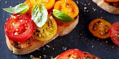 A close-up overhead shot of several pieces of bruschetta topped with sliced heirloom tomatoes, fresh basil leaves, and a sprinkle of coarse salt and pepper, arranged on a dark slate surface.