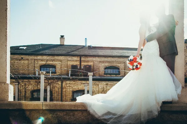 A bride in a white wedding dress holding a colorful bouquet of red and white flowers while standing with a groom in front of a brick building.