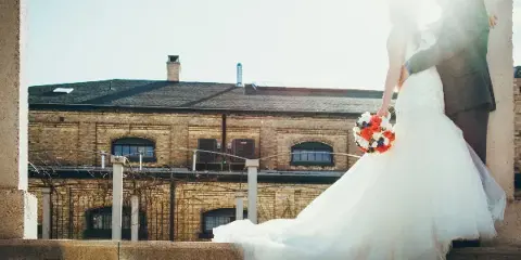 A bride in a white wedding dress holding a colorful bouquet of red and white flowers while standing with a groom in front of a brick building.