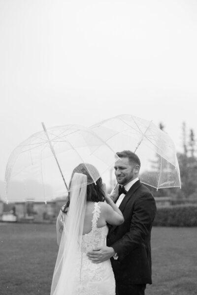A bride and groom embrace under clear umbrellas during a rainy outdoor wedding first look.