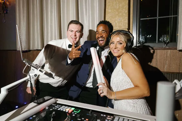 Three wedding entertainment professionals standing behind a DJ mixing console at an event.