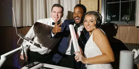 Three wedding entertainment professionals standing behind a DJ mixing console at an event.