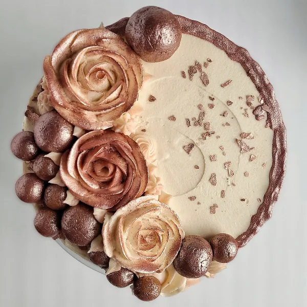 A top-down view of a round cake decorated with piped buttercream roses, chocolate spheres, and gold leaf accents.
