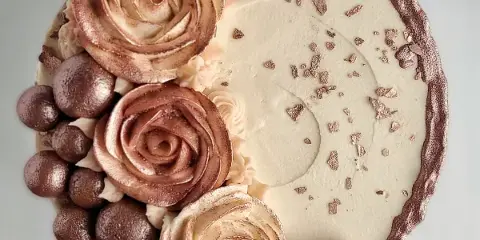 A top-down view of a round cake decorated with piped buttercream roses, chocolate spheres, and gold leaf accents.