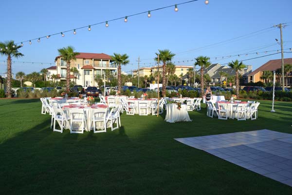 Outdoor event setup with multiple round tables, white chairs, and linens arranged on a lawn with string lights overhead.
