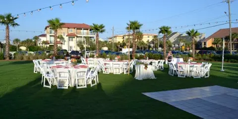 Outdoor event setup with multiple round tables, white chairs, and linens arranged on a lawn with string lights overhead.