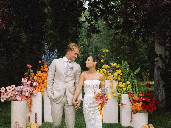 A bride and groom holding hands in front of colorful floral pedestals and greenery.