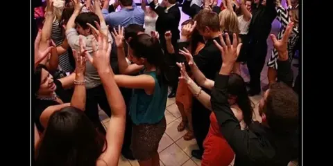 A crowded dance floor at a wedding reception with guests dancing and raising their hands under purple lighting.