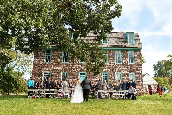 A bride and groom standing at the altar during an outdoor wedding ceremony in front of a large stone building with green window frames.