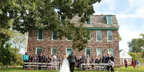 A bride and groom standing at the altar during an outdoor wedding ceremony in front of a large stone building with green window frames.