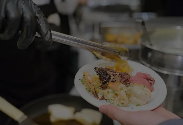 A catering staff member wearing black gloves uses tongs to serve a plate of shrimp and meat.