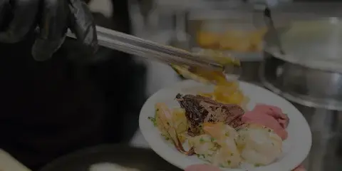 A catering staff member wearing black gloves uses tongs to serve a plate of shrimp and meat.
