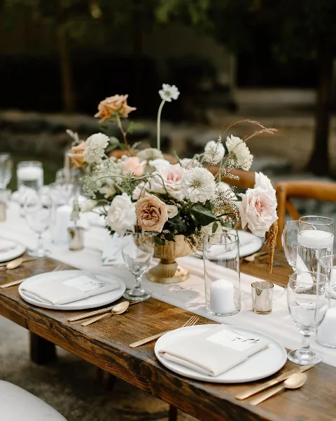 Styled wooden event table set with white plates, glassware, candles, and a floral centerpiece