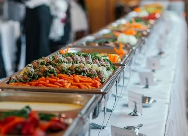 A long catering buffet table with multiple chafing dishes filled with food, garnished with carrots and greens, set with serving spoons and menu cards.