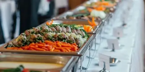 A long catering buffet table with multiple chafing dishes filled with food, garnished with carrots and greens, set with serving spoons and menu cards.
