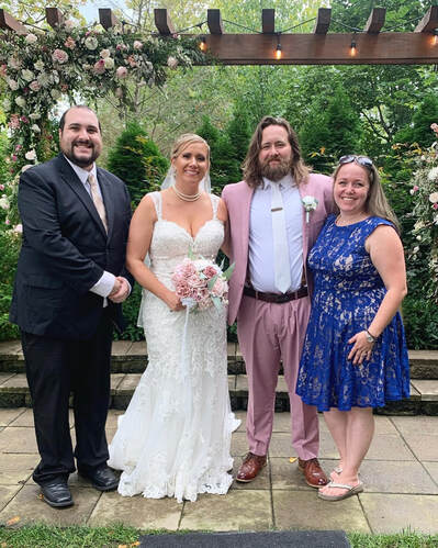 A bride and groom posing with their wedding party in front of a floral-decorated wooden pergola.