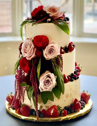 Two-tier white wedding cake decorated with red and pink roses, greenery, and fresh berries on a gold cake board.