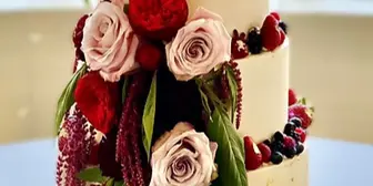 Two-tier white wedding cake decorated with red and pink roses, greenery, and fresh berries on a gold cake board.