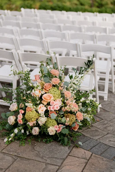 A lush floral arrangement featuring pink roses, hydrangeas, and greenery placed on a stone patio in front of rows of white wedding chairs.