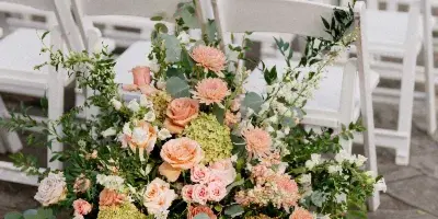 A lush floral arrangement featuring pink roses, hydrangeas, and greenery placed on a stone patio in front of rows of white wedding chairs.