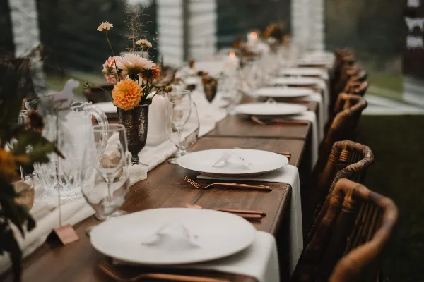 A long wooden farm table set with white plates, glassware, and floral centerpieces, surrounded by rustic wooden chairs.