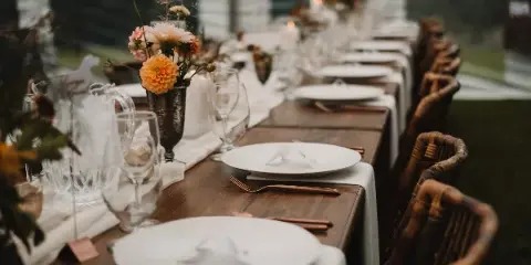 A long wooden farm table set with white plates, glassware, and floral centerpieces, surrounded by rustic wooden chairs.