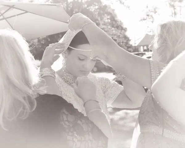 A bride in traditional attire having her headpiece and jewelry adjusted by two women during wedding preparations.