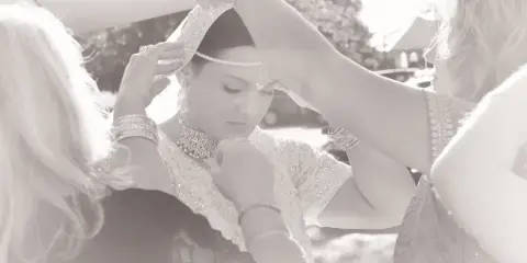 A bride in traditional attire having her headpiece and jewelry adjusted by two women during wedding preparations.