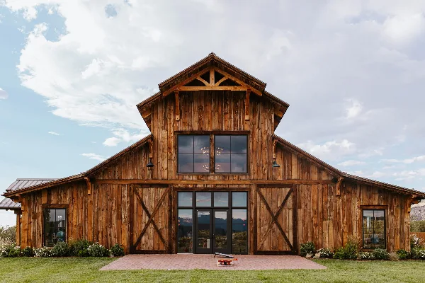 Exterior view of a large modern wooden barn with a steep gabled roof, large glass windows, and a brick entryway, set against a blue sky with clouds.