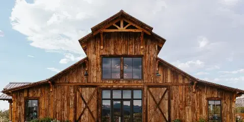 Exterior view of a large modern wooden barn with a steep gabled roof, large glass windows, and a brick entryway, set against a blue sky with clouds.