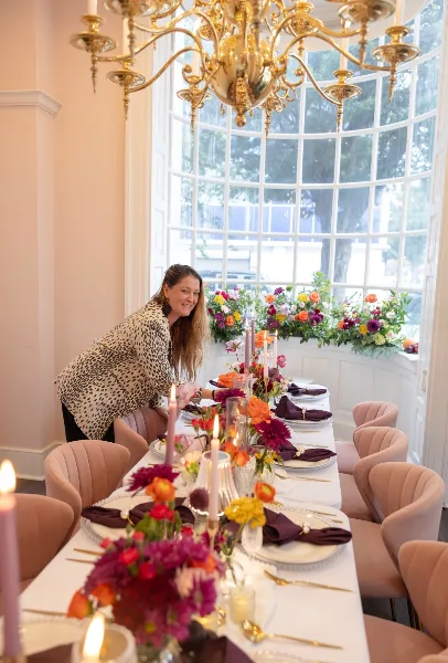 A woman arranging a floral centerpiece on a decorated dining table with candles and place settings.