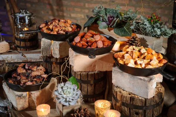 A rustic catering display featuring cast-iron skillets filled with meats and sides, arranged on wooden barrels and stone blocks with floral and candle decorations.