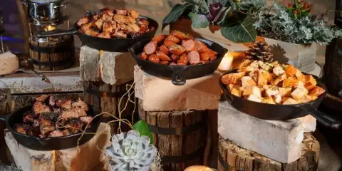 A rustic catering display featuring cast-iron skillets filled with meats and sides, arranged on wooden barrels and stone blocks with floral and candle decorations.